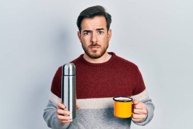 Sakallı, elinde termo kuşkucu ve gergin bir şekilde bir fincan kahve içen yakışıklı beyaz bir adam sorun yüzünden üzgün. negatif kişi. 