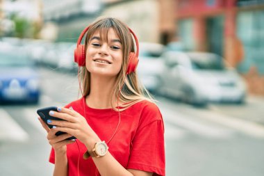 Kafkasyalı genç kız şehirde akıllı telefon ve kulaklık kullanarak mutlu bir şekilde gülümsüyor..