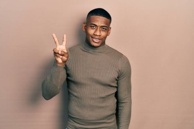 Balıkçı yaka kazak giyen genç siyahi bir adam iki numaralı parmaklarını gösterip gösterirken kendinden emin ve mutlu gülümsüyor.. 