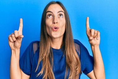 Gündelik giysiler giyen genç ve güzel bir kadın şaşırmış ve şaşırmış bir şekilde yukarı bakıyor ve parmaklarıyla işaret ediyor ve kollarını kaldırıyor.. 