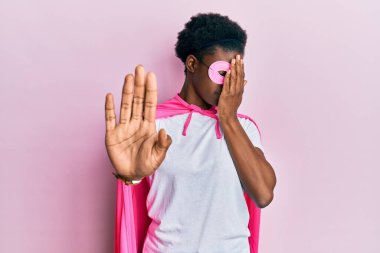 Genç Afro-Amerikalı kız süper kahraman maskesi takıyor ve pelerin giyip gözlerini ellerle kapatıyor ve üzgün ve korku ifadeleriyle jest yapıyor. utangaç ve olumsuz kavram. 