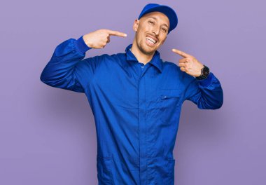 Sakallı, inşaatçı tulumu giyen, neşeli gülümseyen ve parmaklarıyla ve ağzıyla işaret eden kel bir adam. diş sağlığı kavramı. 