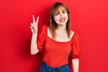 Kırmızı tişörtlü, kızıl saçlı genç bir kadın iki numaralı parmaklarını göstererek gülümseyerek mutlu ve kendinden emin bir şekilde gülümsedi.. 
