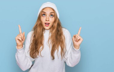 Genç beyaz kız yün kazak ve kış şapkası giyiyor. Şaşırmış ve şaşırmış. Parmaklarıyla işaret ediyor ve kollarını kaldırıyor.. 