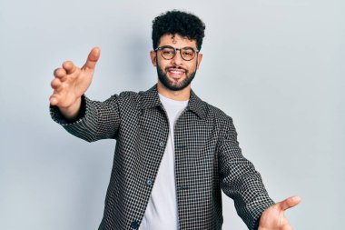 Sakallı, gözlüklü genç bir Arap, kameraya bakıyor. Sarılmak için kollarını açmış gülümsüyor. Mutluluğu kucaklayan neşeli bir ifade.. 