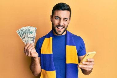 Genç İspanyol futbolcu Amerikan dolarlarını elinde tutan akıllı telefon kullanıyor. Kameraya seksi bir ifadeyle bakıyor, neşeli ve mutlu bir yüzle.. 