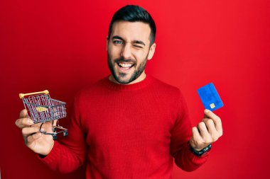 Küçük bir süpermarket arabası ve kredi kartı tutan İspanyol genç adam kameraya seksi bir ifadeyle bakıyor, neşeli ve mutlu bir yüz.. 