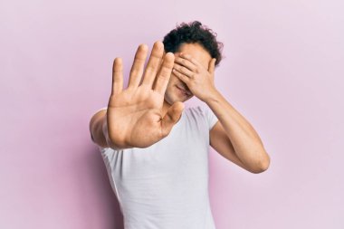 Gündelik beyaz tişört giyen genç yakışıklı adam elleriyle gözlerini kapatıyor ve üzgün ve korku ifadeleriyle jest yapıyor. utangaç ve olumsuz kavram. 