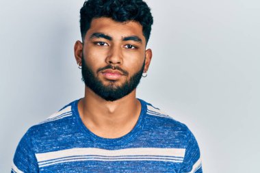 Sakallı, sıradan çizgili tişörtlü, yüzünde ciddi bir ifade olan bir Arap. Basit ve doğal bir şekilde kameraya bakıyorsun.. 