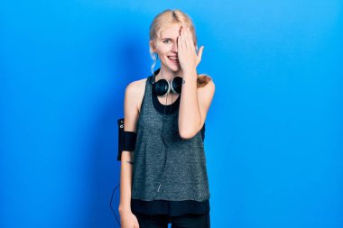 Güzel, beyaz, sarı saçlı, spor giyimli bir kadın. Bir gözünü elinle kapatıyor. Yüzünde kendine güvenen bir gülümseme ve sürpriz bir duygu.. 