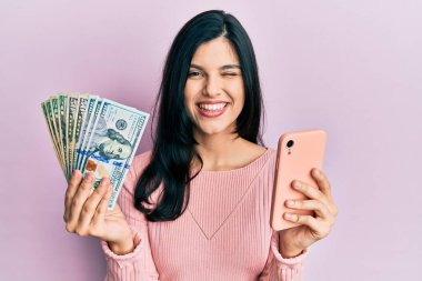 Akıllı telefon kullanan genç İspanyol kadın Amerikan doları banknotları tutuyor. Kameraya seksi bir ifadeyle bakıyor, neşeli ve mutlu bir yüzle.. 