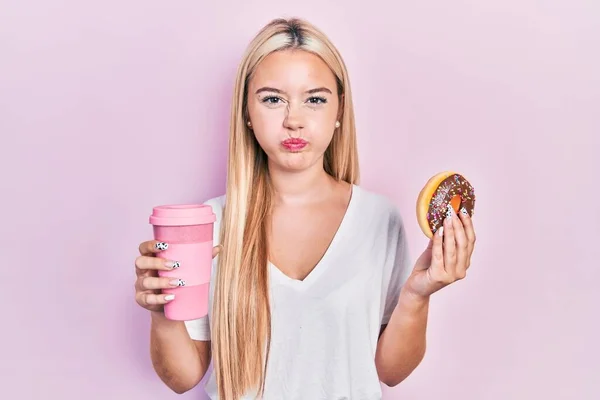 年轻的金发姑娘吃着甜甜圈 喝着咖啡 脸上带着滑稽的表情 嘴被空气吹胀 — 图库照片