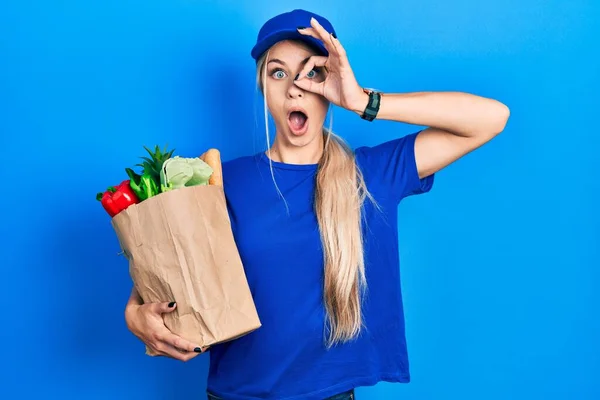 スーパーマーケットからの食料品と宅配便の制服を着た若い白人女性は 驚きの顔でショックを受けたOkジェスチャーを行うと 目を指で見て 信じられない表現 — ストック写真