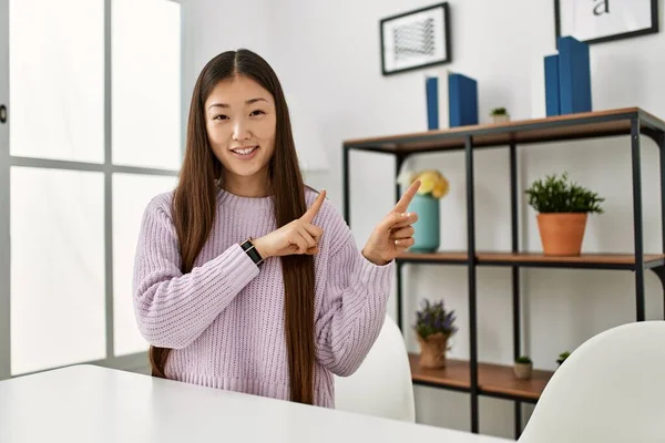 若いです中国の女の子身に着けていますカジュアル服上のテーブルの上に家の笑顔とカメラを見てポインティングで二つの手と指で側面 — ストック写真