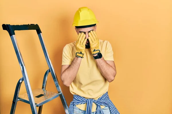 Όμορφος Άντρας Γενειάδα Από Κατασκευαστικές Σκάλες Φορώντας Καπέλο Τρίβοντας Μάτια — Φωτογραφία Αρχείου