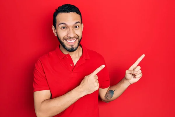 髭を生やしたヒスパニック系の男はカジュアルな赤いTシャツを着て笑顔とカメラを見て2本の手と指で側面を指して — ストック写真