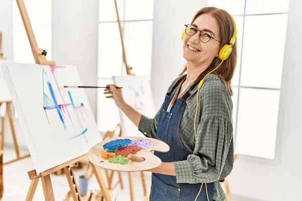 Middle age artist woman smiling happy listening to music and painting at art studio.