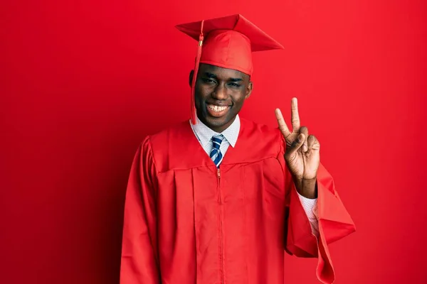 若いアフリカ系アメリカ人の男が卒業キャップと式のローブを着て 自信と幸せな笑顔で指の数2を示して指摘しながら — ストック写真