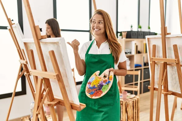 Young hispanic middle age woman at art classroom pointing thumb up to the side smiling happy with open mouth