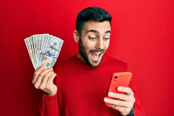 Jovem Hispânico Usando Smartphone Segurando Notas Dólares Dos Eua Sorrindo — Fotografia de Stock