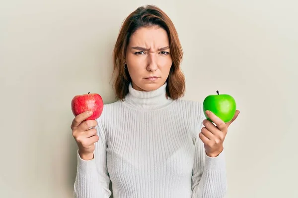 年轻的高加索女人 手里拿着红绿相间的苹果怀疑和紧张 皱着眉头因为问题而烦恼 消极的人 — 图库照片
