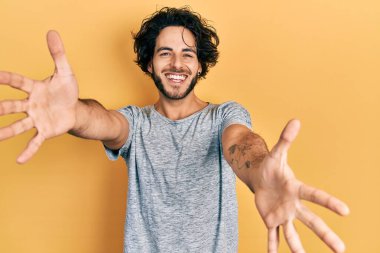 Yakışıklı, gri tişört giyen yakışıklı bir adam kucak açmak için kollarını açmış kameraya bakıyor. Mutluluğu kucaklayan neşeli bir ifade.. 