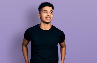 Siyah bir tişört giyen Afro-Amerikan genç bir adam rahat bir görünüm ve kendine güvenen bir gülümseme takınıyor.. 