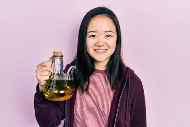 Elinde zeytinyağı tutan genç Çinli kız pozitif ve mutlu görünüyor ve kendine güvenen bir gülümsemeyle dişlerini gösteriyor. 