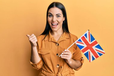 Birleşik krallık bayrağını tutan genç esmer kadın başparmağını yana kaldırmış, ağzı açık gülümsüyor. 