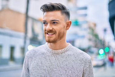 Genç beyaz adam gülümseyerek şehirde mutlu mesut duruyor..