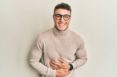 Balıkçı yaka kazak giyen İspanyol genç adam gülümseyerek kahkahalarla gülüyor çünkü elleri vücutta olan komik bir şaka.. 