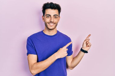 Gündelik tişört giyen genç İspanyol adam gülümseyip kameraya bakıyor ve iki eliyle ve parmaklarıyla kenarı işaret ediyor.. 