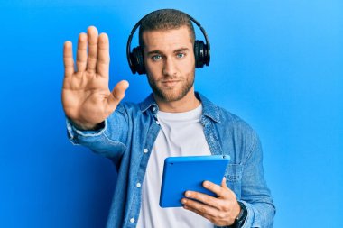 Genç beyaz adam dokunmatik ped kullanıyor kulaklık takıyor ve eli açık dur işareti yapıyor ciddi ve kendinden emin bir ifadeyle, savunma hareketi yapıyor. 