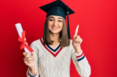 Mezuniyet diploması olan genç esmer kız bir fikirle gülümsüyor ya da mutlu bir yüzle işaret ediyor. 