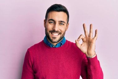 Gündelik kıyafetler giyen genç İspanyol adam gülümsüyor. El ve parmaklarıyla 