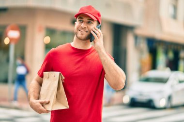 Beyaz atlı genç teslimatçı akıllı telefondan konuşuyor ve şehirde kağıt torba taşıyor..