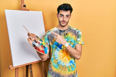 İspanyol asıllı genç adam paletle resim çiziyor, resim sehpası ayakta duruyor, el parmağı yanda duruyor, ciddi ve sakin bir yüz ifadesi var. 