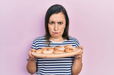 Orta yaşlı İspanyol kadın elinde tatlı pastalar tutarken şüpheci ve gergin, sorun yüzünden surat asıyor. negatif kişi. 