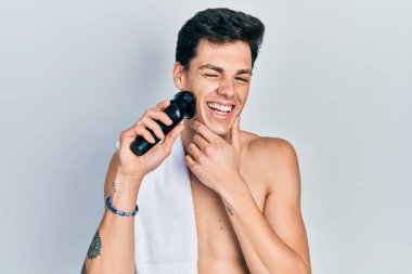 Elektrikli tıraş makinesiyle tıraş olan genç İspanyol adam kameraya seksi bir ifadeyle bakıyor, neşeli ve mutlu bir yüz.. 
