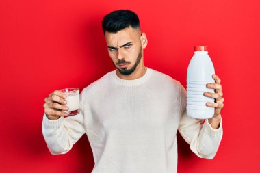 Sakallı, elinde bir bardak süt ve plastik şişe tutan, kuşkucu ve gergin bir İspanyol genç, sorun yüzünden surat asıyor. negatif kişi. 