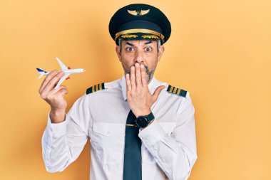 Yakışıklı, orta yaşlı, gri saçlı, uçak pilotu üniforması giyen, oyuncak uçak tutan, ağzı elle kaplanmış, şok olmuş ve hata yapmaktan korkan bir adam. şaşırmış ifade 