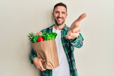 Sakallı yakışıklı bir adam, elinde poşetlerle, güler yüzlü, dostça bir selamlaşma ve selamlaşma sunuyor. başarılı iş. 