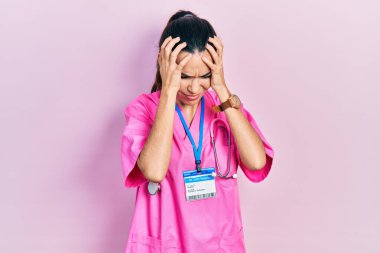 Genç esmer kadın doktor üniforması giyiyor ve steteskop baş ağrısı çekiyor. Acı ve migren yüzünden çaresiz ve stresli. Eller başının üstünde.. 