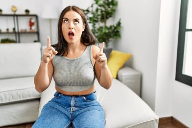 Evdeki kanepede oturan genç esmer kadın şaşırmış ve şaşırmış bir şekilde yukarı baktı ve parmaklarını ve kollarını kaldırdı.. 