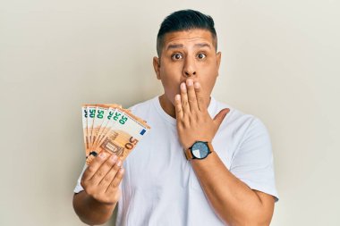Elinde 50 avroluk banknotlar tutan genç latin adam ağızlarını eliyle kapatıyor, şaşırmış ve hata yapmaktan korkuyor. şaşırmış ifade 