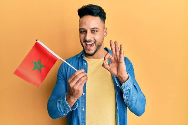 Fas bayrağı tutan genç Arap adam parmaklarıyla imza atıyor, gülümseyerek mükemmel bir sembol oluşturuyor. 