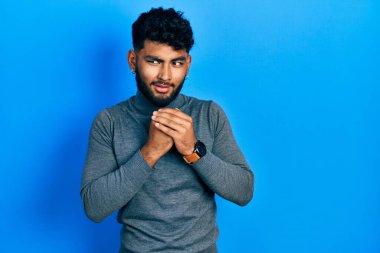 Balıkçı yaka kazak giyen sakallı bir Arap elleri çenesinde gergin ve heyecanlı bir şekilde yan tarafa bakıyor. 