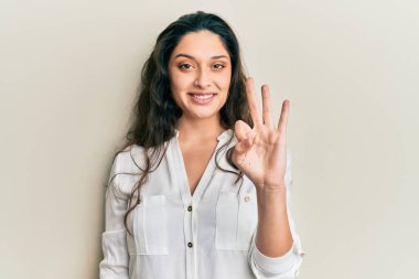 Ortadoğulu güzel bir kadın gündelik kıyafetler giyer ve üç numaralı parmaklarıyla gülümseyerek mutlu ve kendinden emin bir şekilde onu işaret eder.. 