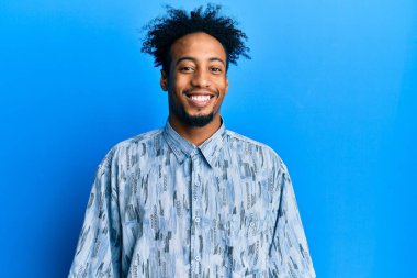 Sakallı, sıradan kıyafetler giyen, mutlu ve havalı bir gülümsemesi olan Afro-Amerikalı genç bir adam. şanslı kişi. 