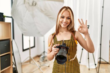 Kafkasyalı genç fotoğrafçı kız elinde profesyonel kamerayla fotoğraf stüdyosunda tamam işareti yapıyor, gülümseyerek mükemmel bir sembol oluşturuyor. 